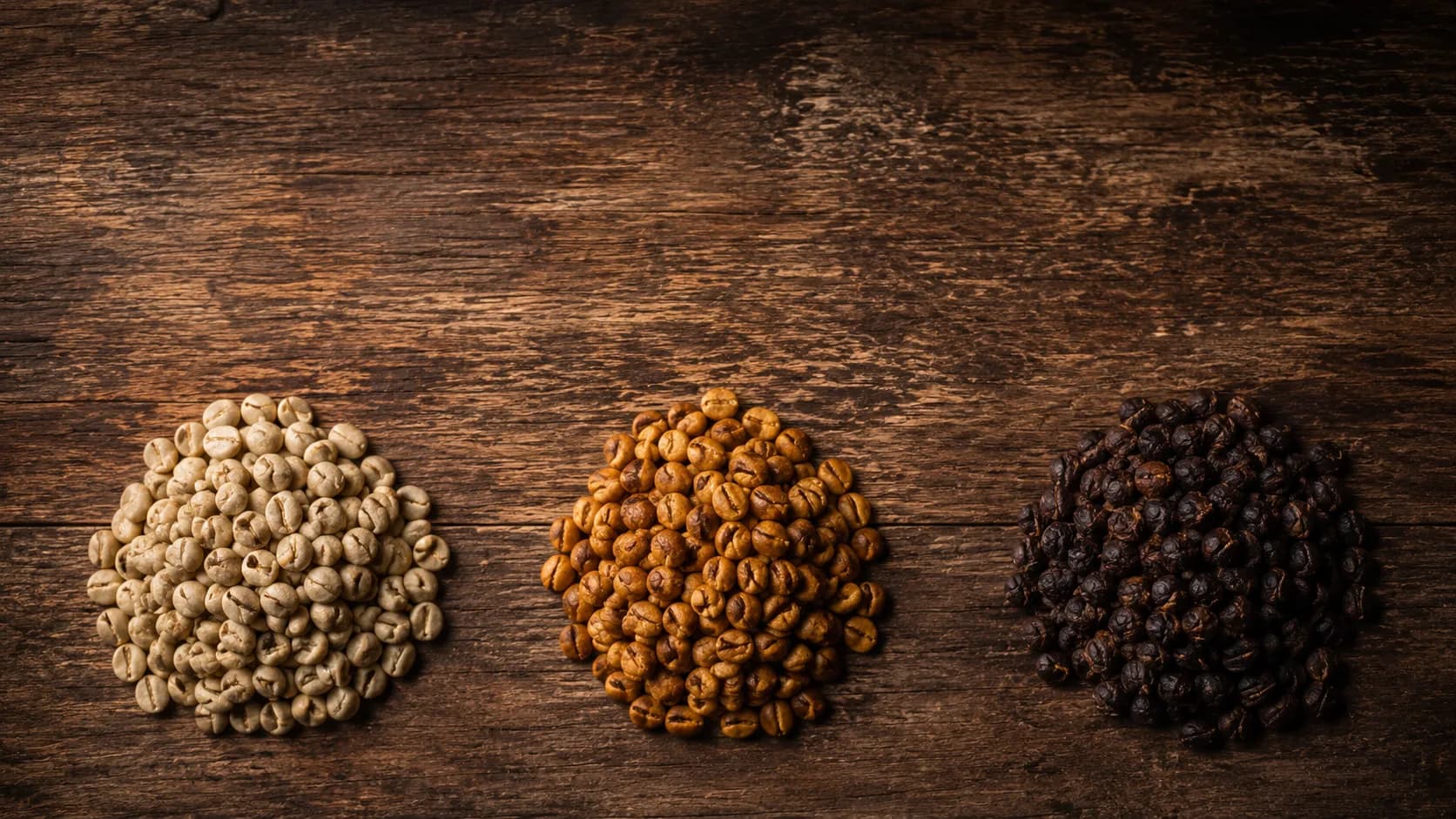 Tres montones de granos de café —crudo, tostado medio y oscuro— sobre madera.